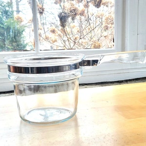 May include: A clear glass saucepan with a transparent handle and a silver-colored metal band around the middle. The saucepan is sitting on a wooden surface near a window.