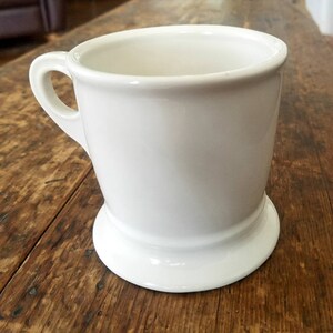 May include: A white ceramic shaving mug with a wide base and a handle. The mug is empty and ready to be used.