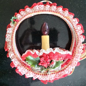 May include: A circular wall decoration featuring a central faux candle with a dark bulb, surrounded by layers of white and red lace trim. The base is adorned with green holly leaves, red berries, and small silver bells, creating a festive display.
