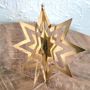 May include: A gold-coloured, multi-layered star ornament. The star has a three-dimensional design with multiple points and cut-out sections. The ornament is likely for holiday or seasonal decor.