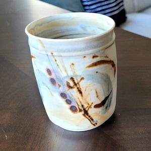 May include: A white ceramic mug with a brown, blue, and purple abstract design. The mug has a rounded shape and a slightly flared lip.
