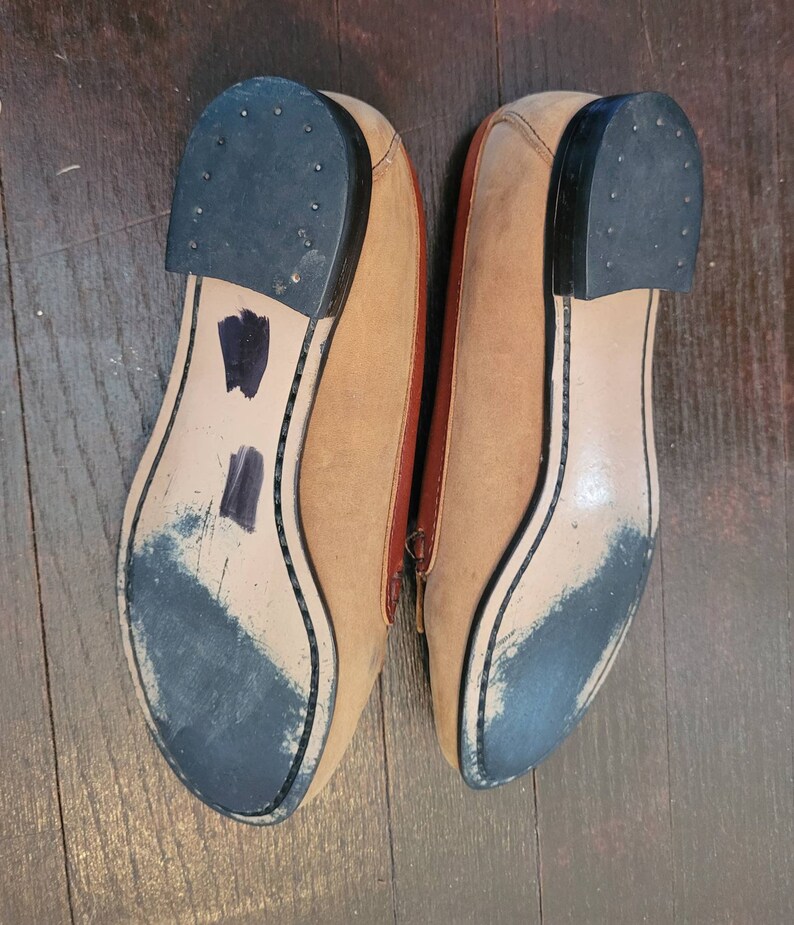 May include: A pair of tan leather loafers with black soles. The loafers have a brown leather trim around the edges.