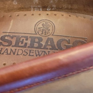 May include: Close-up of the inside of a brown leather shoe with the brand name "Sebago" and the text "Handsewn" printed on the insole. A small logo of a man sitting on a chair is visible above the brand name.