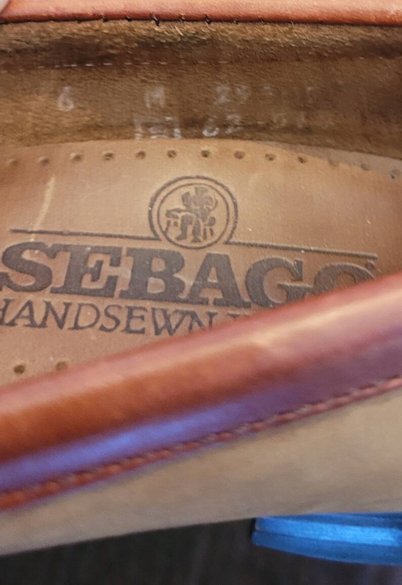May include: Close-up of the inside of a brown leather shoe with the brand name "Sebago" and the text "Handsewn" printed on the insole. A small logo of a man sitting on a chair is visible above the brand name.