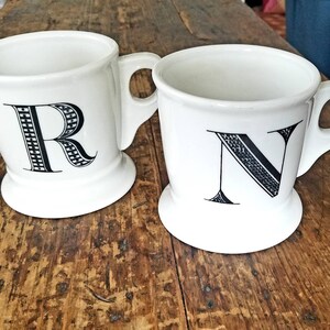 May include: Two white ceramic mugs with black letter designs. The mug on the left has the letter "R" and the mug on the right has the letter "N".