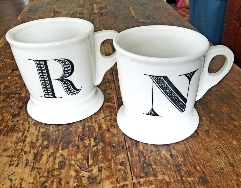 May include: Two white ceramic mugs with black letter designs. The mug on the left has the letter "R" and the mug on the right has the letter "N".