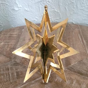 May include: A gold-coloured, layered star ornament with a central star design. The ornament has a three-dimensional effect, with multiple star layers. The star is suspended from a small loop at the top, suggesting it is designed for hanging.