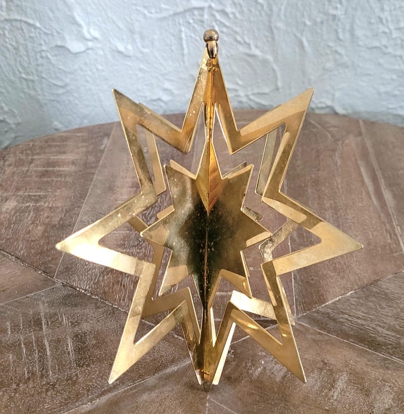 May include: A gold-coloured, layered star ornament with a central star design. The ornament has a three-dimensional effect, with multiple star layers. The star is suspended from a small loop at the top, suggesting it is designed for hanging.