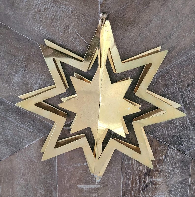 May include: A decorative gold-coloured star ornament. The star is made of layered metal sheets, creating a three-dimensional effect. It has multiple points and hangs from a small, round, silver-coloured ball.