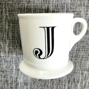 May include: White ceramic mug with a black letter J on the front. The mug has a wide base and a handle.