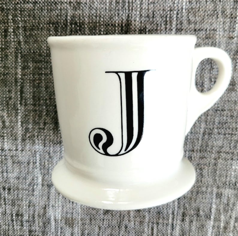 May include: White ceramic mug with a black letter J on the front. The mug has a wide base and a handle.