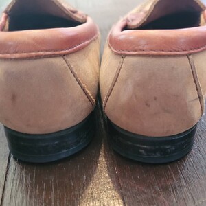May include: A pair of brown suede leather loafers with a brown leather trim around the top. The loafers have black rubber soles.