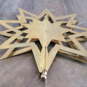 May include: A gold-coloured star-shaped ornament with multiple points and a layered design. The star is made of metal and has a matte finish. The ornament is likely for holiday or seasonal decor.