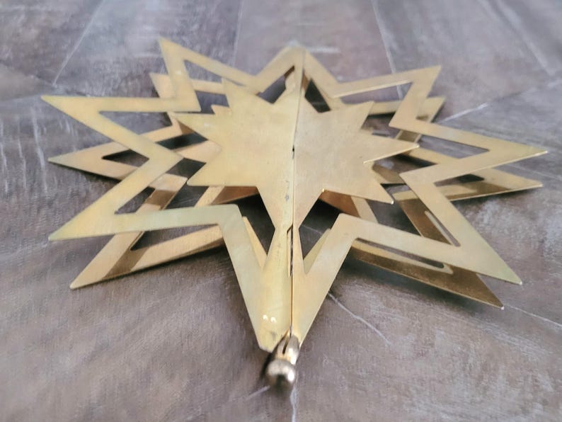 May include: A gold-coloured star-shaped ornament with multiple points and a layered design. The star is made of metal and has a matte finish. The ornament is likely for holiday or seasonal decor.