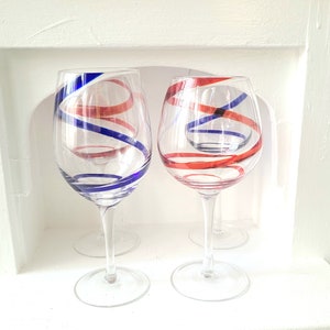 May include: Two clear wine glasses with red and blue spiral designs. The glasses have long stems and sit on a white surface. The spiral designs are painted on the outside of the glasses, creating a decorative effect.