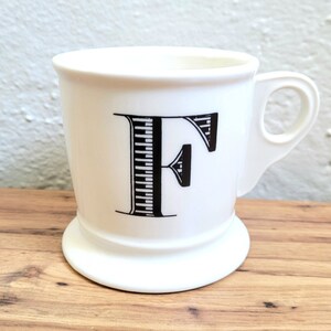May include: White ceramic mug with a black and white striped letter F on the front. The mug has a handle and a wide base.