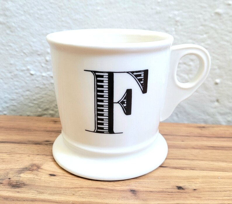 May include: White ceramic mug with a black and white striped letter F on the front. The mug has a handle and a wide base.