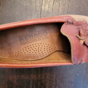 May include: Close-up of the inside of a brown leather loafer with a red leather trim and a fringe detail. The insole is brown leather with the text "SEBAGO HANDSEWN IN U.S.A." printed on it.