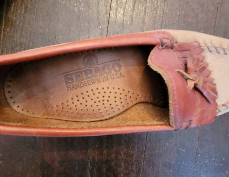 May include: Close-up of the inside of a brown leather loafer with a red leather trim and a fringe detail. The insole is brown leather with the text "SEBAGO HANDSEWN IN U.S.A." printed on it.