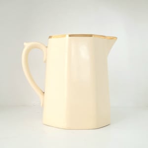 May include: A cream-colored ceramic pitcher with a gold rim. The pitcher has an octagonal shape and a handle.