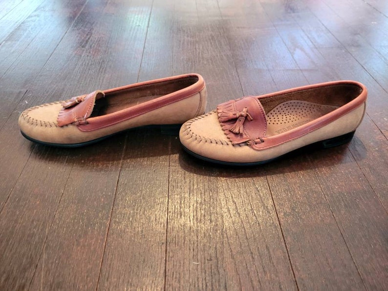 May include: A pair of brown leather loafers with tassels. The loafers have a light brown upper and a darker brown trim around the edges. The tassels are made of the same leather as the loafers.