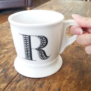 May include: White ceramic mug with a black and white patterned letter "R" on the front. The mug has a handle and a wide base.