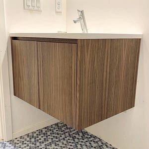 May include: A brown wood vanity with a white countertop and a chrome faucet. The vanity has three doors and is mounted on the wall above a hexagonal tile floor.