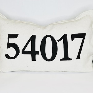 Könnte beinhalten: Ein weißes Kissen mit schwarzen gestickten Zahlen 54017.