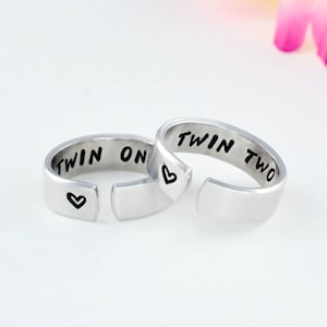 May include: Two silver rings with the words "Twin One" and "Twin Two" engraved on them. Each ring has a small heart symbol engraved on the side.