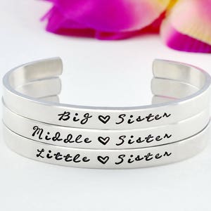 May include: Three silver bangle bracelets with the words "Big Sister", "Middle Sister", and "Little Sister" engraved on them. Each bracelet has a heart symbol between the words "Sister" and the word before it.