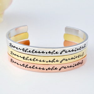 May include: Three stacked bracelets, each with the inscription "Nevertheless, she persisted" in a different metal finish: silver, gold, and rose gold.