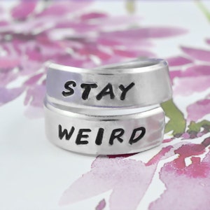 May include: A silver ring with the words "STAY WEIRD" engraved on it. The ring is a simple band with a slightly curved design.
