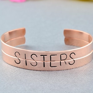 May include: Rose gold double band bangle bracelet with the word "SISTERS" engraved on the front.