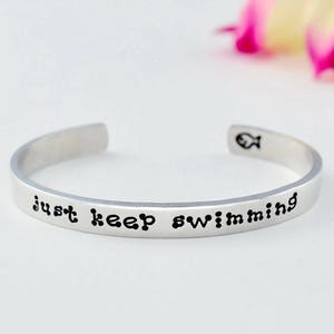 May include: Silver bracelet with the words "just keep swimming" engraved on it. The bracelet has a small fish charm.