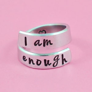 May include: A silver ring with the words "I am enough" engraved on it. The ring is open and can be adjusted to fit different finger sizes.