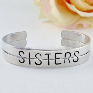 May include: A silver-colored cuff bracelet with the word "SISTERS" engraved in black block letters. The bracelet has a simple, modern design and is open at the ends.