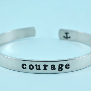 May include: A silver-colored cuff bracelet with the word "courage" stamped in black, centered on the front. An anchor symbol is stamped on one end. The bracelet is open-ended and sits against a light blue background.