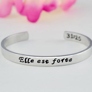 May include: Silver bangle bracelet with the inscription "Elle est forte" in black lettering. The bracelet has a 31/25 stamp on the back.