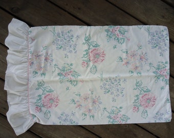 May include: White pillowcase with a floral pattern featuring pink and purple flowers with green leaves. The pillowcase has a ruffled edge with pink stitching.