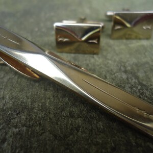 May include: A gold-toned tie clip and matching cufflinks set. The tie clip has a geometric design with a subtle etched pattern. The cufflinks are rectangular with a similar design. The set is displayed on a textured, green surface.