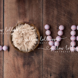 Puede incluir: La palabra "LOVE" deletreada con pelotas de béisbol rosas y blancas sobre un fondo de madera marrón. Un pequeño nido beige y esponjoso se encuentra en la esquina superior izquierda de la imagen.