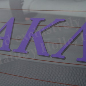 May include: Purple vinyl decal with the letters "AKA" on a window. The letters are in a bold, sans-serif font. The decal is likely for a car or other vehicle. The background is a light gray with red lines.