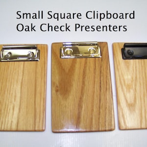 May include: Three rectangular oak wood clipboards with metal clips, labeled Small Square Clipboard Oak Check Presenters. Two clipboards feature shiny silver-toned metal clips, while the third clipboard on the right has a matte black metal clip.