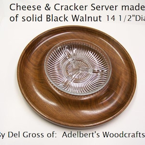 May include: A round wooden cheese and cracker server made of solid black walnut. The server is 14 1/2 inches in diameter and features a glass dish with three compartments in the center. The text "Cheese & Cracker Server made of solid Black Walnut 14 1/2"Dia. By Del Gross of: Adelbert's Woodcrafts" is visible in the image.