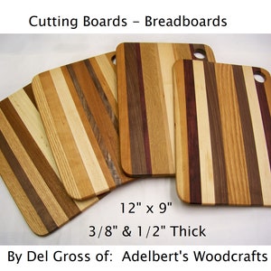 May include: Four wooden cutting boards, each with a different wood grain pattern, are arranged in a staggered formation. The boards are 12 inches by 9 inches and are 3/8 inch and 1/2 inch thick. The text "Cutting Boards - Breadboards" is at the top of the image. The text "By Del Gross of: Adelbert's Woodcrafts" is at the bottom of the image.