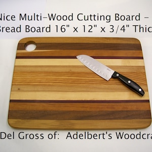 May include: A multi-wood cutting board with a hole in the top. The board is 16 inches by 12 inches and 3/4 inch thick. It is made of different types of wood, including maple, cherry, and walnut. A knife is lying on the board.