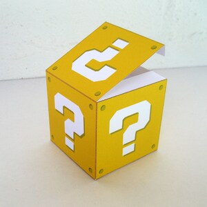 Könnte beinhalten: Eine gelbe Pappbox in Form eines Fragezeichenblocks aus dem Videospiel Super Mario Bros. Die Box hat einen Klappdeckel, der geöffnet ist und den Innenraum der Box freigibt.