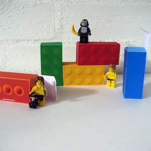 May include: A set of colorful Lego-style building blocks made of paper. The blocks are red, yellow, green, and blue. There are also three small figurines: a monkey, a man with a spear, and a man with a loincloth. The text "paper download" is printed on one of the blocks.