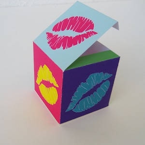 Puede incluir: Una pequeña caja colorida con una tapa con bisagras. La caja está decorada con cuatro labios diferentes, cada uno de un color diferente: rosa, amarillo, turquesa y verde.