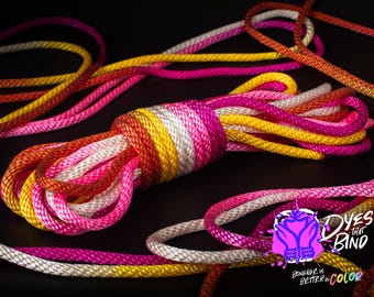 May include: A close-up of a multi-colored rope made of white, yellow, pink, and orange strands. The rope is coiled and arranged in a decorative pattern. The text "Dyes That Bind" is visible in the lower right corner of the image.
